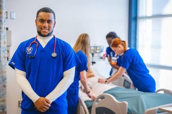 Shenandoah University nursing students, wearing blue scrubs, gathered around a manikin (simulated patient) in a bed.